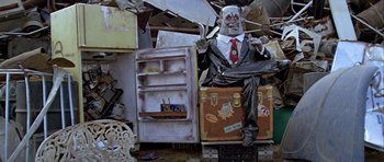 Movie still from “Heartbeeps” (1981), directed by Allan Arkush – A statue of a man in a suit sitting on top of a suitcase next to an open refrigerator; Wide shot, Low angle