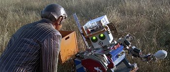 Movie still from “Heartbeeps” (1981), directed by Allan Arkush – A man standing next to a robot in a grassy field; Medium shot, High angle