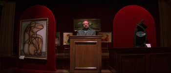 Movie still from “Heartbreakers” (2001), directed by David Mirkin – A man standing at a podium in front of paintings; Wide shot, Low angle
