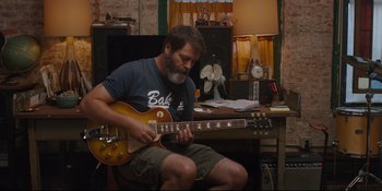 Movie still from “Hearts Beat Loud” (2018), directed by Brett Haley – A man sitting at a desk playing a guitar; Medium shot, Over the shoulder angle