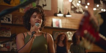 Movie still from “Hearts Beat Loud” (2018), directed by Brett Haley – A woman holding a microphone in front of a group of people; Medium shot, Over the shoulder angle