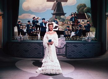 Movie still from “Hello Frisco, Hello” (1943), directed by H. Bruce Humberstone – A woman in a white dress standing in front of a painting of a windmill; Wide shot, Low angle