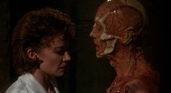 Movie still from “Hellraiser” (1987), directed by Clive Barker – A woman standing next to a man with a fake face; Close Up shot, Over the shoulder angle
