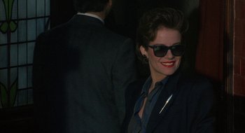 Movie still from “Hellraiser” (1987), directed by Clive Barker – A woman in a suit and sunglasses smiling for the camera; Close Up shot, Over the shoulder angle