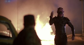 Movie still from “Hellraiser III: Hell on Earth” (1992), directed by Anthony Hickox – A person wearing a demon mask standing in front of a fire; Medium shot, Over the shoulder angle