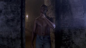 Movie still from “Hellraiser: Deader” (2005), directed by Rick Bota – A shirtless man standing in a dark room with his hand to his mouth; Medium shot, Low angle