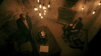 Movie still from “Hellraiser: Judgment” (2018), directed by Gary J. Tunnicliffe – Two people sitting at a table in a dimly - lit room; Wide shot, High angle