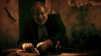 Movie still from “Hellraiser: Judgment” (2018), directed by Gary J. Tunnicliffe – An older man sitting at a table with a bottle; Medium shot, Over the shoulder angle
