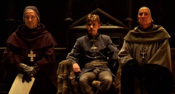 Movie still from “Henry V” (1989), directed by Kenneth Branagh – A man sitting on a chair in front of a wooden wall; Medium shot, High angle