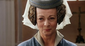 Movie still from “Henry V” (1989), directed by Kenneth Branagh – An older woman wearing a hat and looking at the camera; Close Up shot, Over the shoulder angle