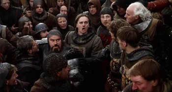 Movie still from “Henry V” (1989), directed by Kenneth Branagh – A group of men in medieval clothing gathered around a man holding a helmet; Medium shot, High angle