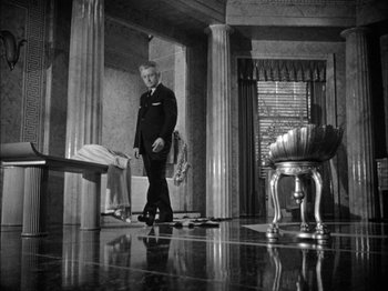 Movie still from “Here Comes Mr. Jordan” (1941), directed by Alexander Hall – A man in a suit and tie standing in front of a table; Wide shot, Low angle