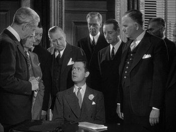 Movie still from “Here Comes Mr. Jordan” (1941), directed by Alexander Hall – A group of men standing around a man in a suit; Medium shot, Over the shoulder angle