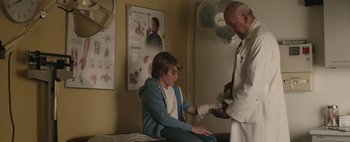 Movie still from “Hesher” (2010), directed by Spencer Susser – A man in a doctor's office is giving a boy an injection in his hand; Medium shot, Low angle