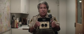 Movie still from “Hesher” (2010), directed by Spencer Susser – A police officer holding up an old style camera; Close Up shot, High angle