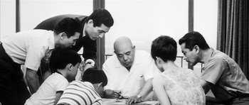 Movie still from “High and Low” (1963), directed by Akira Kurosawa – A group of people gathered around a man writing on a piece of paper; Medium shot, Low angle