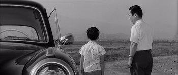 Movie still from “High and Low” (1963), directed by Akira Kurosawa – A young boy standing in front of an old car; Medium shot, Over the shoulder angle