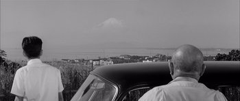 Movie still from “High and Low” (1963), directed by Akira Kurosawa – A man standing next to an old car with a mountain in the background; Medium shot, High angle