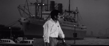 Movie still from “High and Low” (1963), directed by Akira Kurosawa – A black and white photo of a man sitting on a bench in front of a ship; Medium shot, Low angle