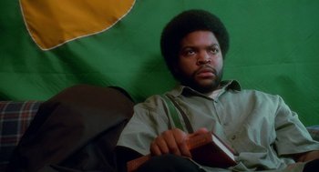 Movie still from “Higher Learning” (1995), directed by John Singleton – A man sitting on a couch holding a book in his hands; Medium shot, Low angle