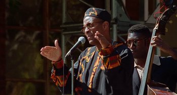 Movie still from “Higher Learning” (1995), directed by John Singleton – A man in a black and orange shirt is speaking into a microphone; Medium shot, Low angle