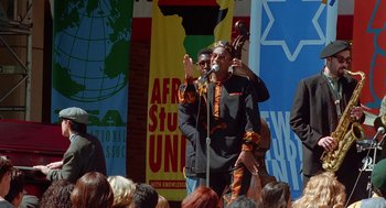Movie still from “Higher Learning” (1995), directed by John Singleton – A man is speaking into a microphone in front of a group of onlookers; Wide shot, High angle
