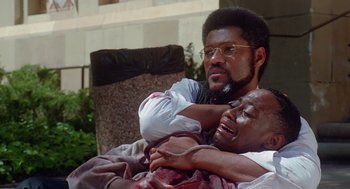 Movie still from “Higher Learning” (1995), directed by John Singleton – A man holding another man's arm while sitting on a couch; Medium shot, High angle