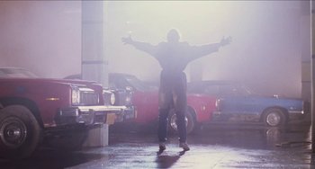 Movie still from “Highlander” (1986), directed by Russell Mulcahy – A man standing on the ground with his arms outstretched in front of cars; Wide shot, Low angle