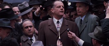 Movie still from “Hoffa” (1992), directed by Danny DeVito – A man in a brown suit is being interviewed by reporters; Medium shot, Over the shoulder angle