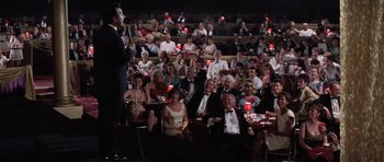 Movie still from “Hoffa” (1992), directed by Danny DeVito – An audience of people sitting at tables and chairs in front of a microphone; Wide shot, High angle