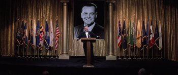 Movie still from “Hoffa” (1992), directed by Danny DeVito – A man standing at a podium in front of a portrait of a man; Medium shot, Low angle