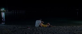 Movie still from “Holiday” (2018), directed by Isabella Eklöf – A man sitting on the ground next to another man on the beach at night; Wide shot, High angle