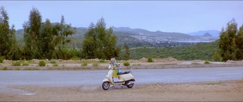 Movie still from “Holiday” (2018), directed by Isabella Eklöf – A person riding a scooter down a dirt road near trees; Extreme Wide shot, High angle