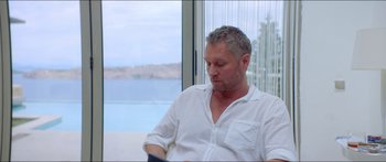 Movie still from “Holiday” (2018), directed by Isabella Eklöf – A man sitting in front of a window looking at something; Medium shot, Over the shoulder angle