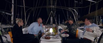 Movie still from “Holiday” (2018), directed by Isabella Eklöf – Two men sitting at a table on top of a sailboat at night; Wide shot, Over the shoulder angle