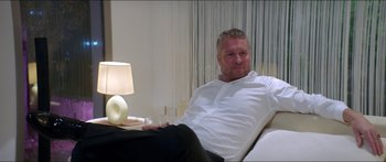 Movie still from “Holiday” (2018), directed by Isabella Eklöf – A man sitting on top of a couch next to a lamp; Medium shot, Over the shoulder angle