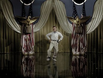 Movie still from “Holiday Inn” (1942), directed by Robert Allen – A man standing in front of american flags and an eagle statue; Wide shot, High angle