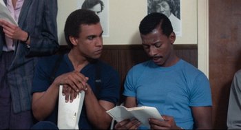 Movie still from “Hollywood Shuffle” (1987), directed by Robert Townsend – A couple of men sitting next to each other holding papers; Medium shot, Over the shoulder angle