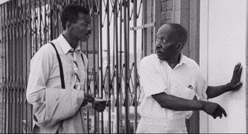 Movie still from “Hollywood Shuffle” (1987), directed by Robert Townsend – Two men standing next to each other in front of a gate; Medium shot, Low angle