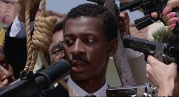 Movie still from “Hollywood Shuffle” (1987), directed by Robert Townsend – A man is speaking into a microphone in front of a group of people; Close Up shot, Low angle