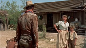 Movie still from “Hondo” (1953), directed by John Farrow – A man and a woman standing in front of a log cabin; Wide shot, Over the shoulder angle