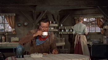 Movie still from “Hondo” (1953), directed by John Farrow – A man drinking a cup of coffee while sitting at a table; Medium shot, Low angle