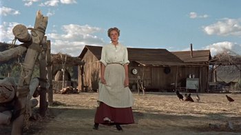 Movie still from “Hondo” (1953), directed by John Farrow – An old woman standing in front of a wooden house; Wide shot, Low angle
