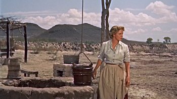 Movie still from “Hondo” (1953), directed by John Farrow – A woman standing next to a bucket in a field; Wide shot, Low angle