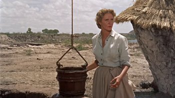 Movie still from “Hondo” (1953), directed by John Farrow – A woman standing next to a bucket on top of the ground; Medium shot, Low angle