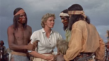 Movie still from “Hondo” (1953), directed by John Farrow – A woman and a group of men standing next to each other on a beach; Medium shot, Over the shoulder angle