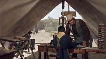 Movie still from “Hondo” (1953), directed by John Farrow – Two men sitting at a wooden table in an old western setting; Wide shot, Over the shoulder angle
