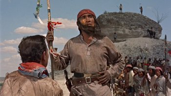Movie still from “Hondo” (1953), directed by John Farrow – A man in a pirate costume holding a flag in front of a group of people; Medium shot, Over the shoulder angle