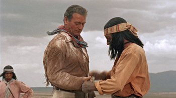 Movie still from “Hondo” (1953), directed by John Farrow – Two men in native american garb , one of them tying the wrist of another man; Medium shot, Over the shoulder angle