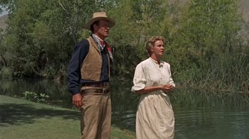 Movie still from “Hondo” (1953), directed by John Farrow – A man and a woman standing next to a body of water; Wide shot, Low angle